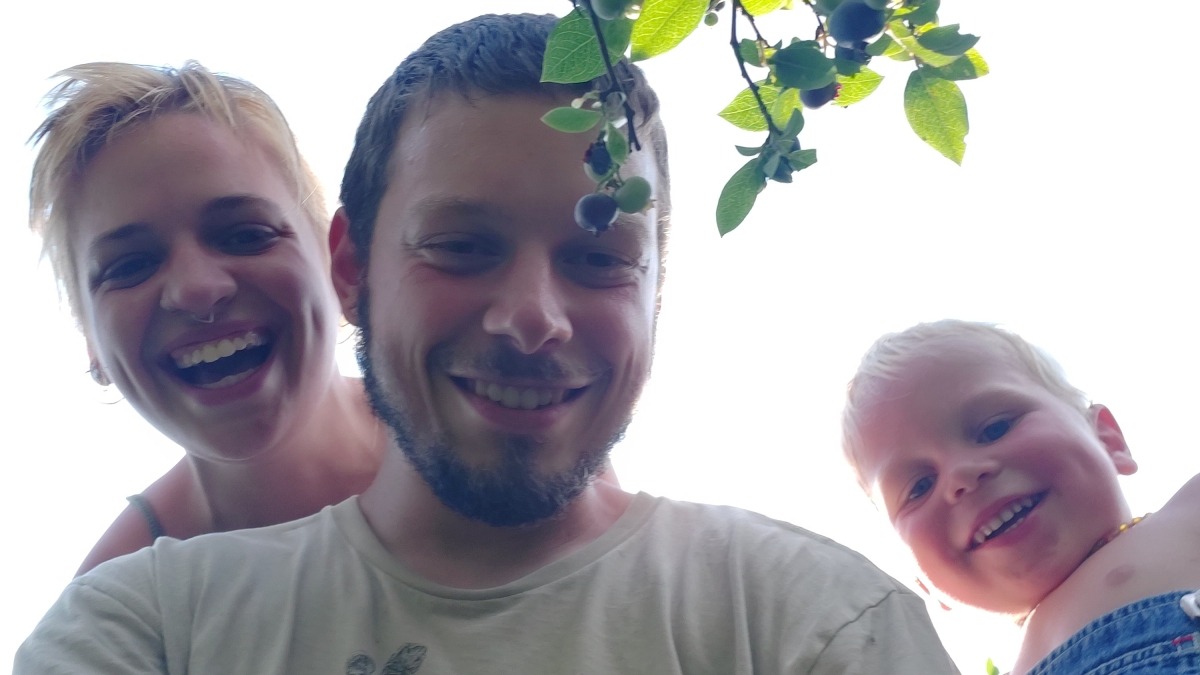 A family of three looking at the camera which is pointed up from the ground with a young woman on the left, a young man in the middle, and a boy toddler on the right all smiling