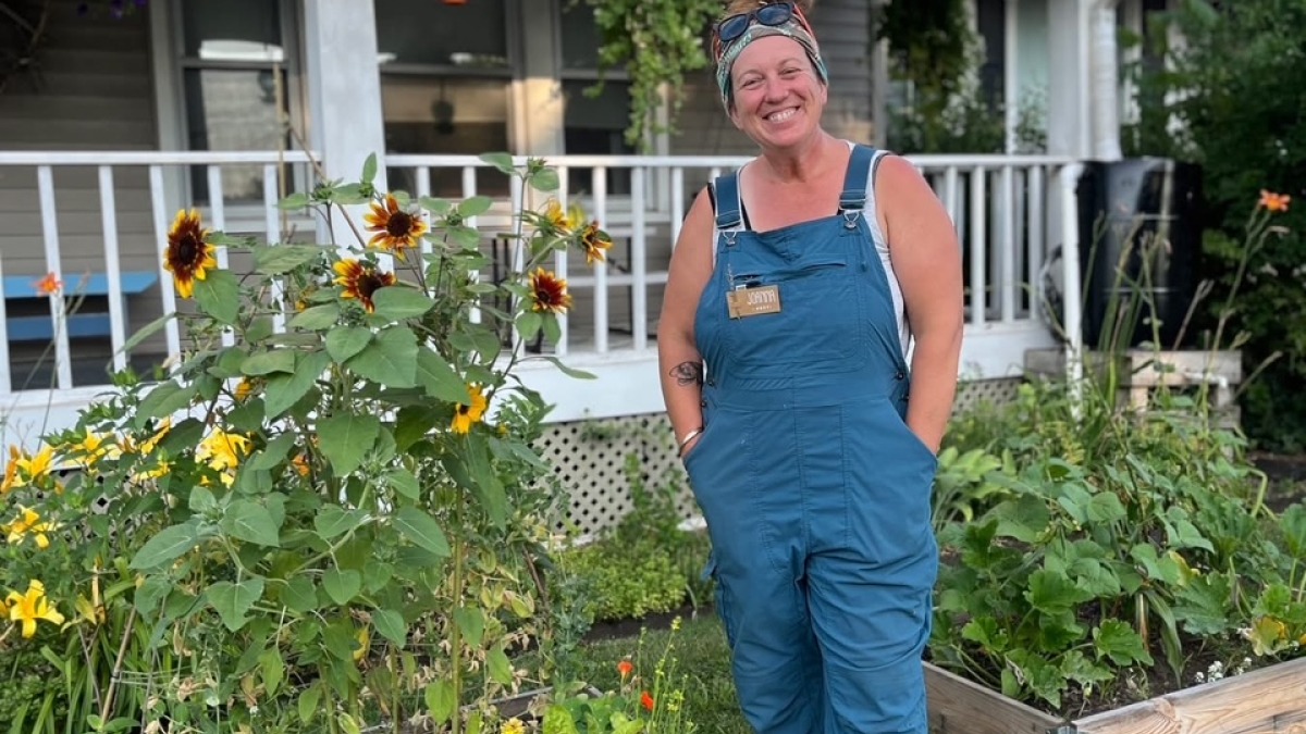 A woman in overalls stands in front of two raised beds full of sunflowers and vegetables. Raised beds are in front of a house in the city with a porch supporting plants and bird feeders.