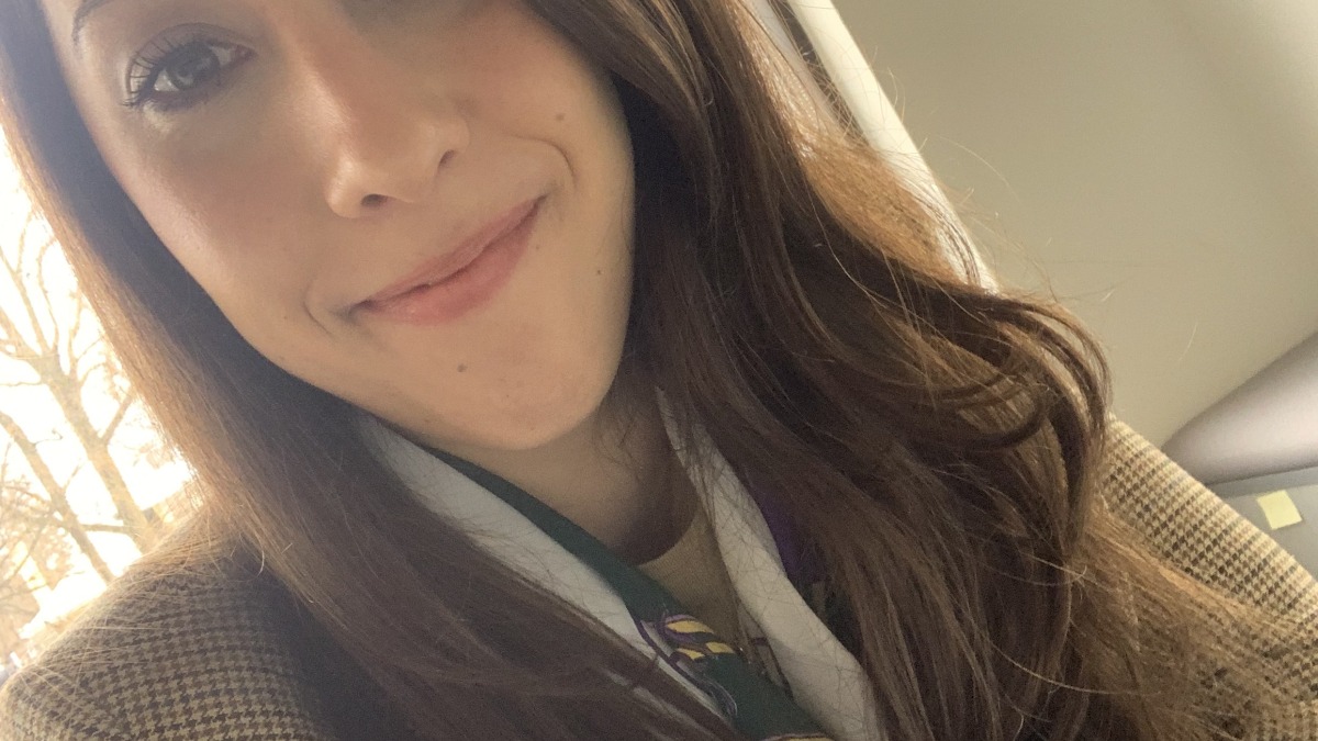 woman with long brunette hair wearing a tan coat and scarf, photographed indoors with natural window light
