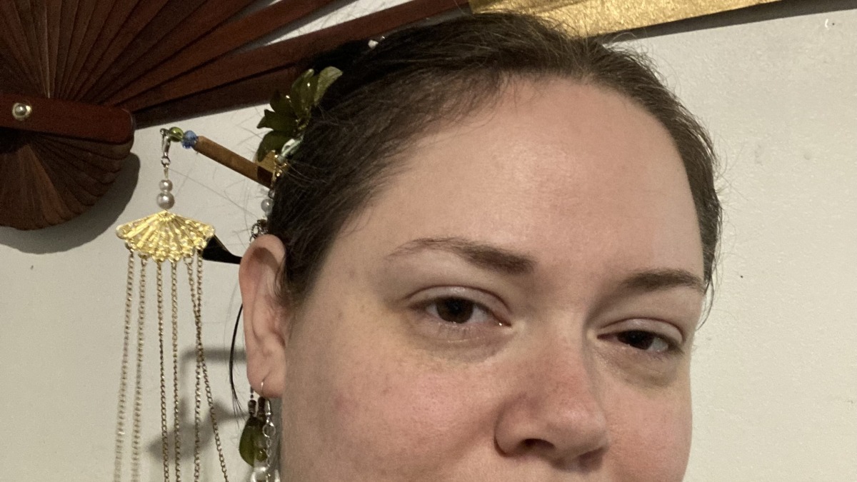 A feminine presenting person with a large ornate folding fan hung on the wall behind them, smiling, with their hair pulled back and various hair ornaments worn, some that dangle, others that do not.