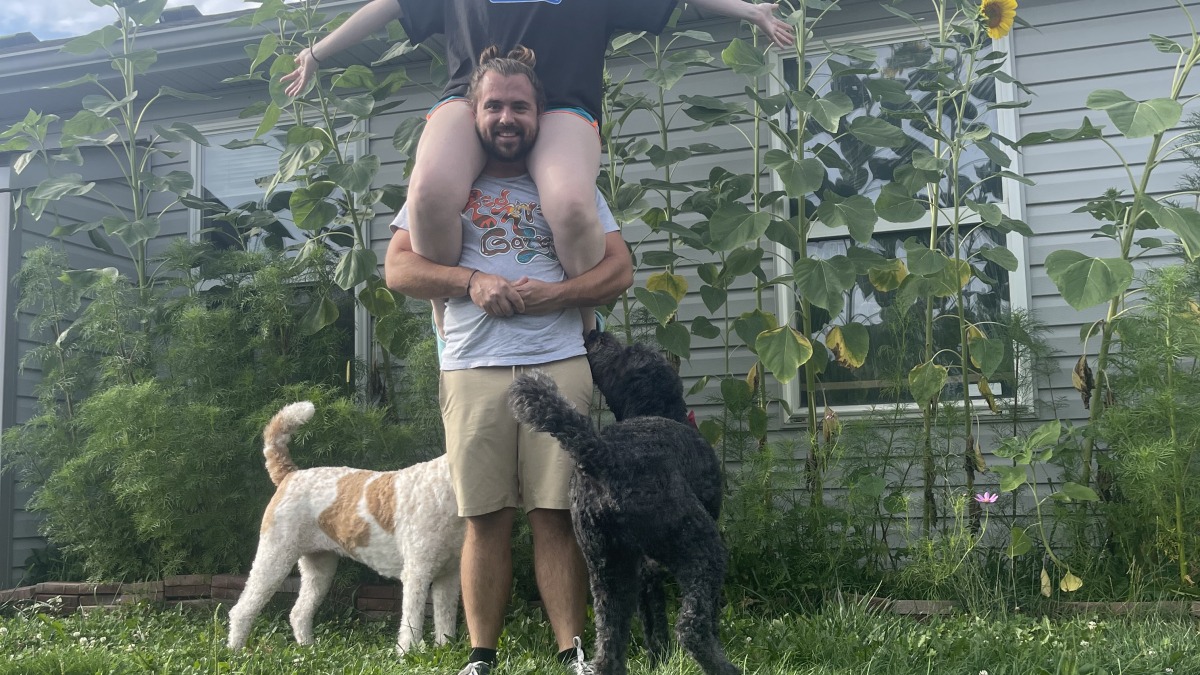 A man hoisting his wife up on his shoulders. Two dogs are in the foreground with a row of sunflowers in the back. 