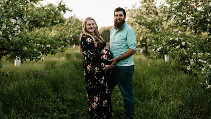 Husband and pregnant wife smiling in an orchard.